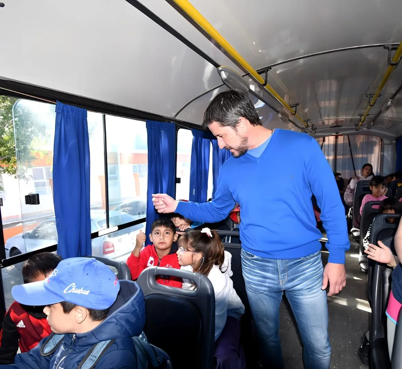 A man in a blue sweater interacts with children seated on a school bus, as daylight streams through large windows, highlighting the students' engaged expressions.