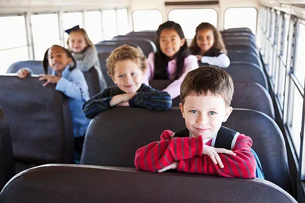 A group of happy children sitting in seats on a school bus, with a focus on a smiling boy in a red striped shirt at the front, embodying the excitement of a school trip or daily commute.