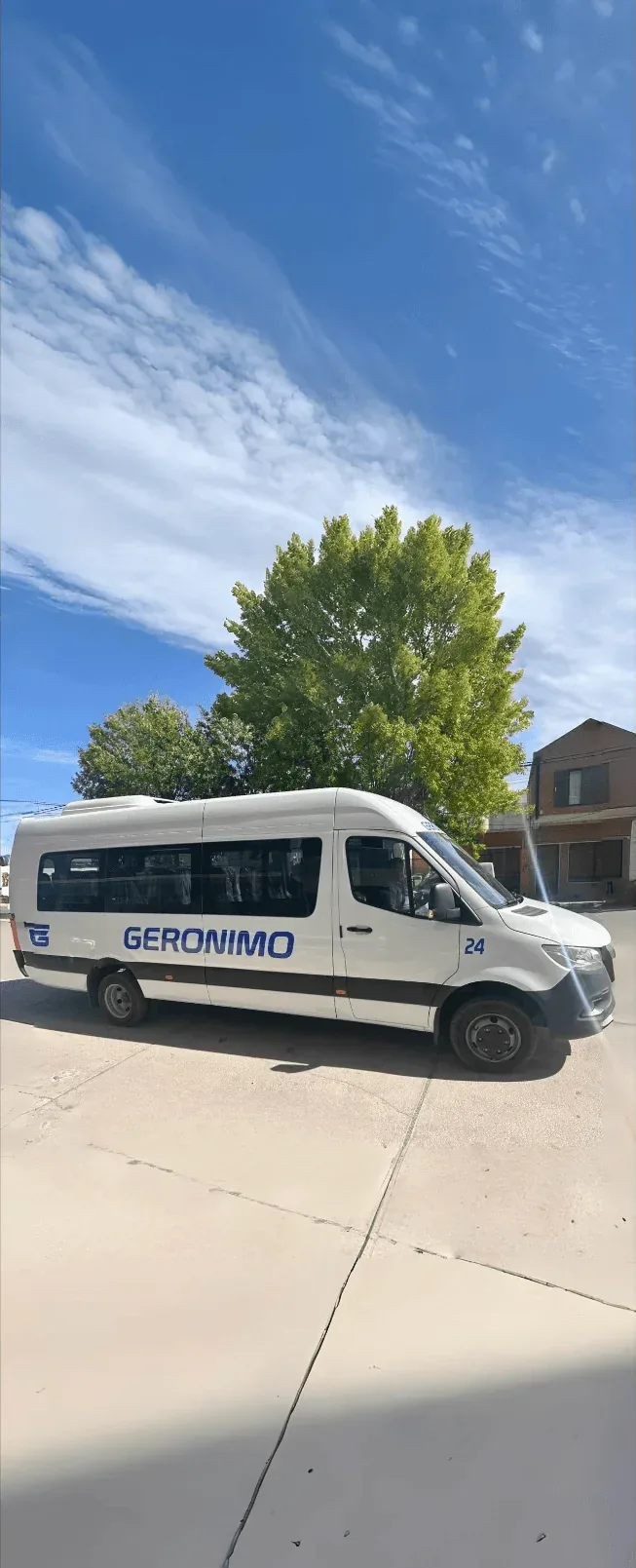A white passenger van with the word "GERONIMO" in blue font is parked on a sunny street beside a large green tree and a brick house.
