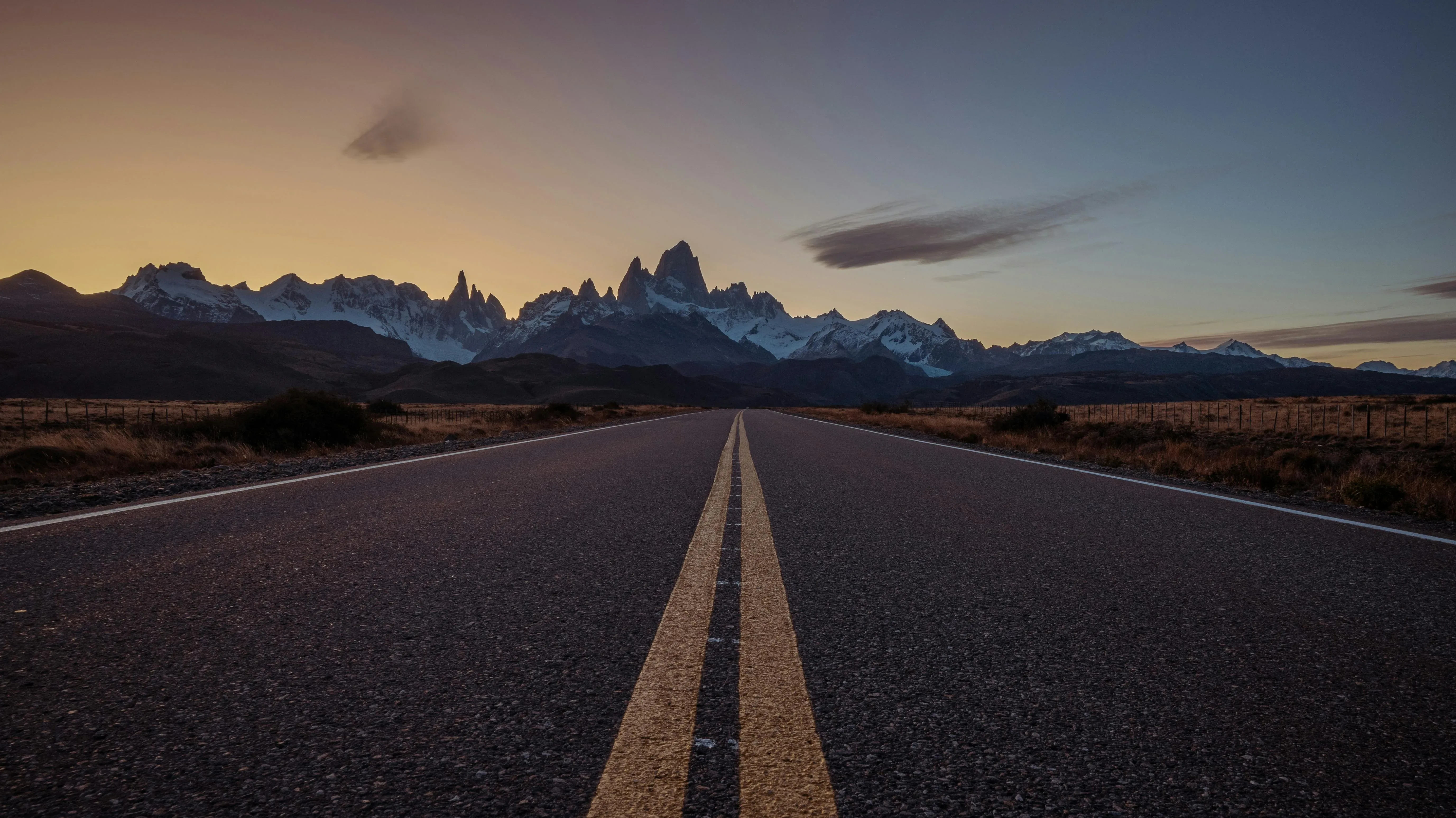 A straight road leads towards a breathtaking mountain range at sunset, with a lavender and orange sky creating a stunning backdrop in Patagonia.