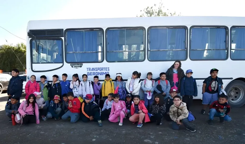 A group of children wearing colorful clothing are gathered in front of a white bus with large windows and blue curtains, parked on a gravel surface.