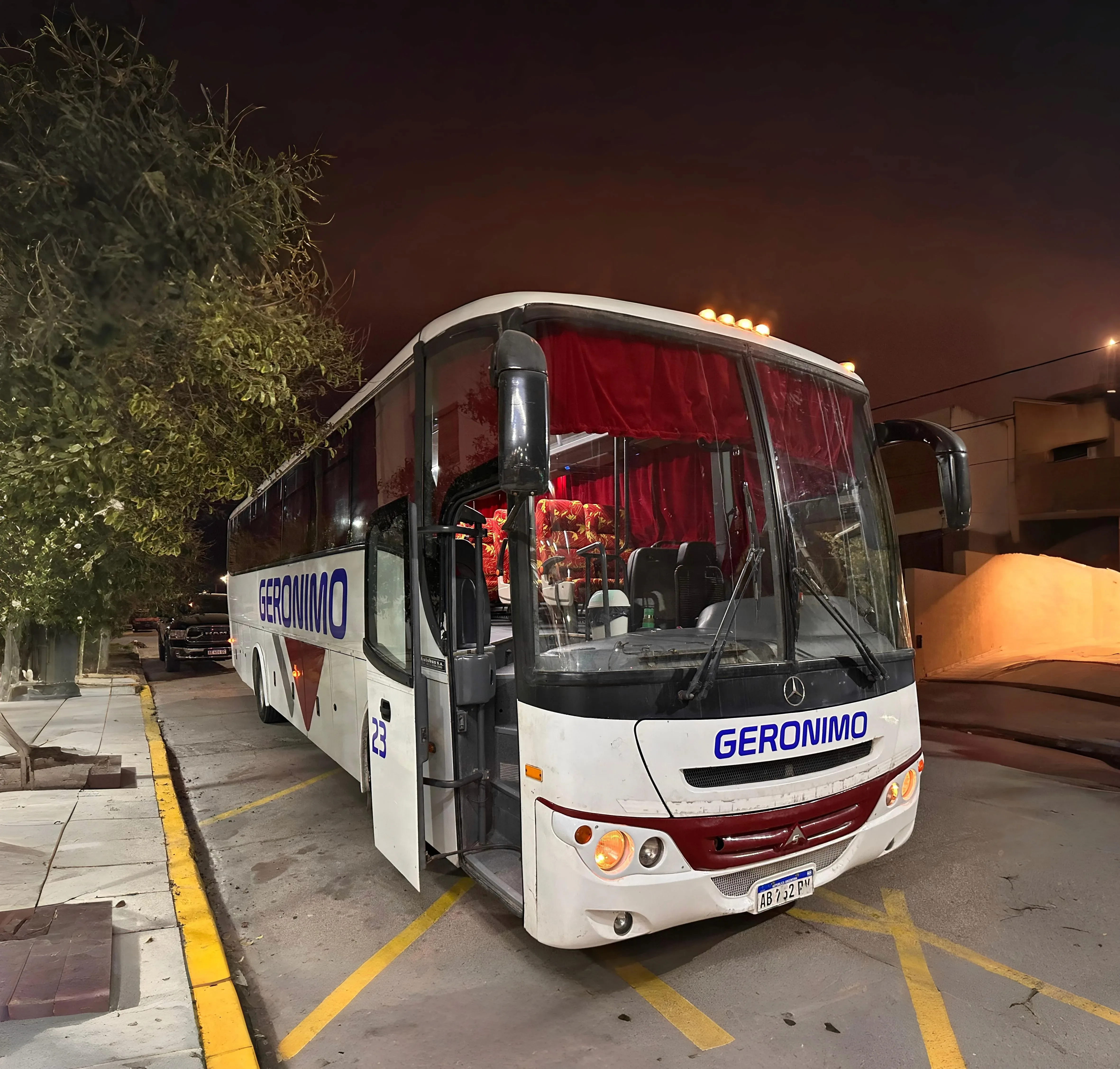 A white bus with "GERONIMO" written on the side is parked on a dimly lit city street, with surrounding trees and sandy terrain visible at night.