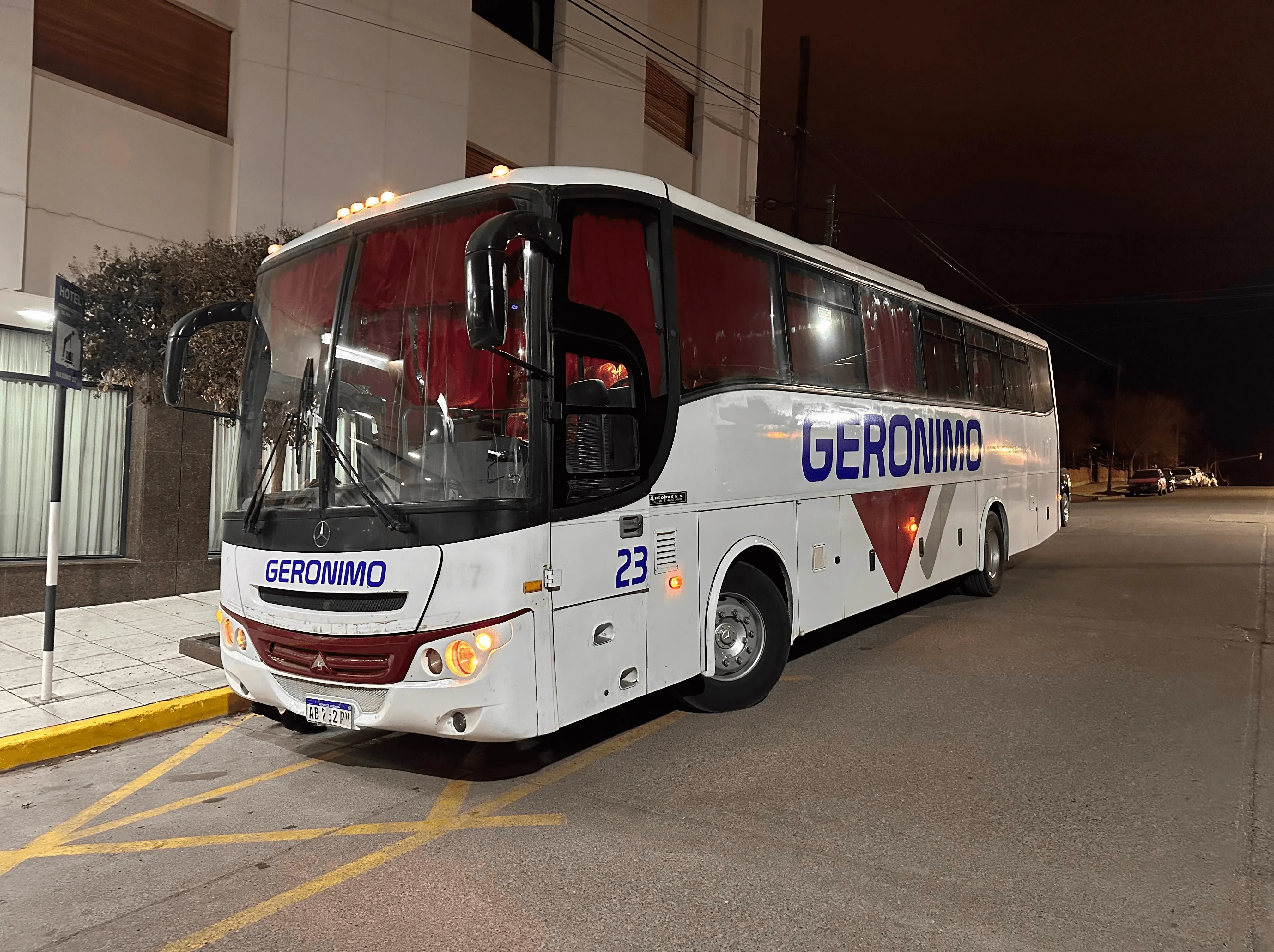 A large white passenger bus with the name "Geronimo" and number "23" on its sides is parked on a well-lit street outside a modern building at night.