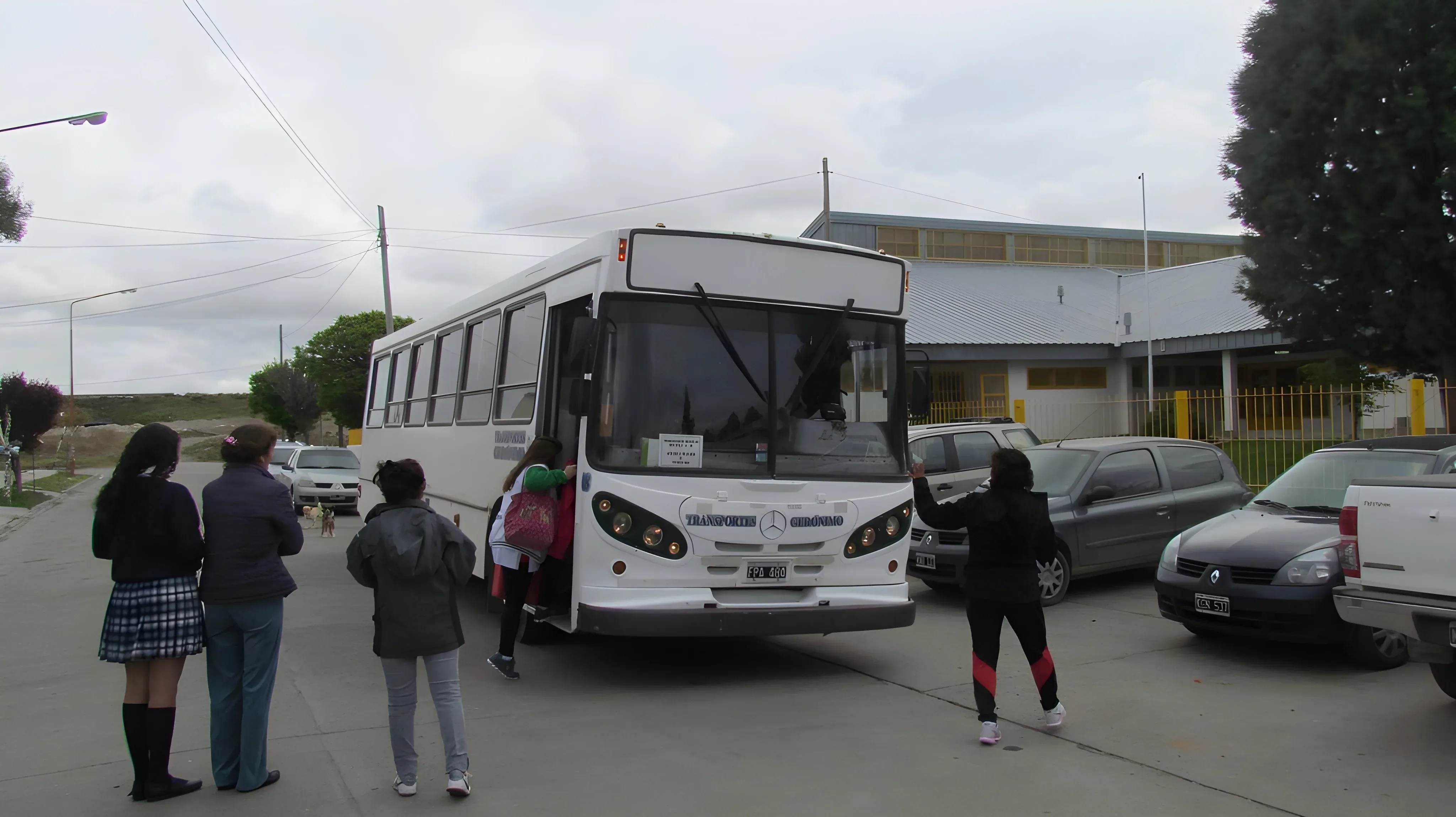 A group of students gathers around a white bus labeled "Transporte Escolar" parked in a school parking lot on a cloudy day.