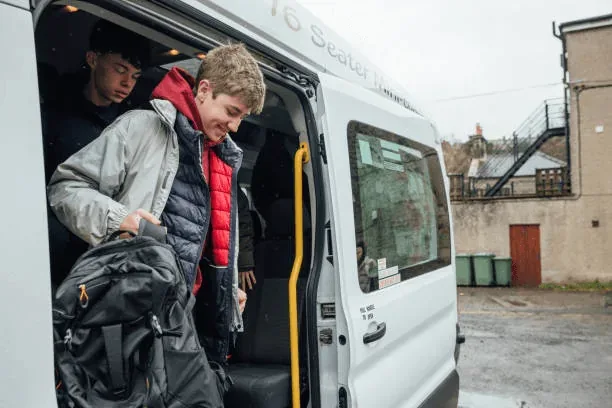 Two people exit a white 16-seater van parked on a wet street, one holding a black backpack, dressed in casual jackets with buildings visible in the background.