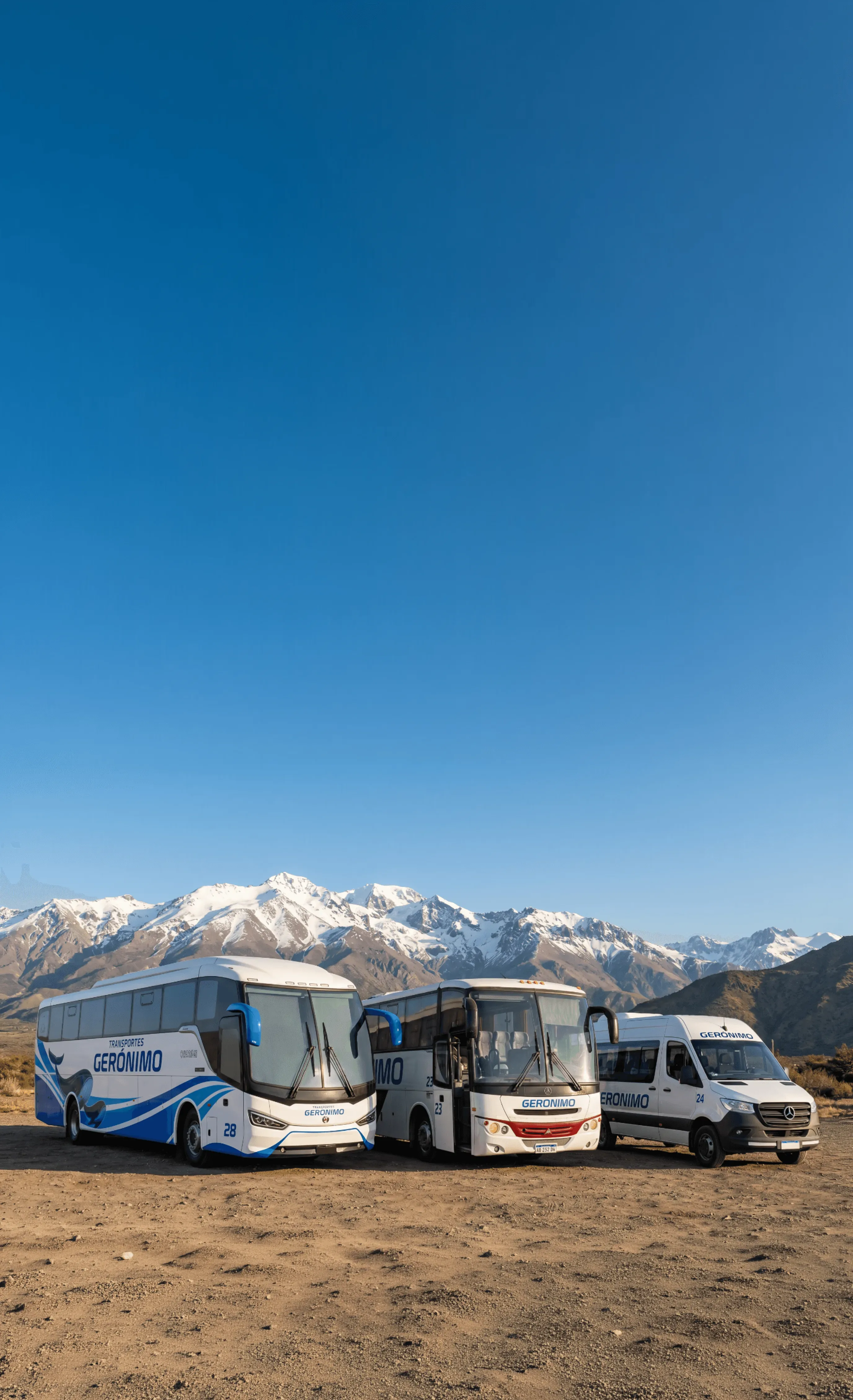 A white tour bus travels on a paved road beside towering rocky cliffs under a clear blue sky with scattered clouds.