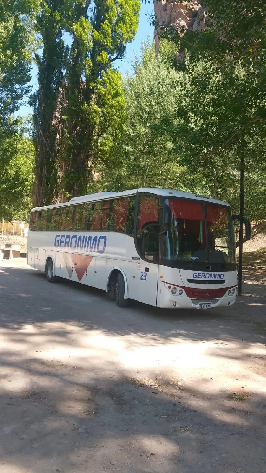 A white tour bus with "Geronimo" written on the side is parked on a dirt road surrounded by tall, lush green trees, under clear blue skies.