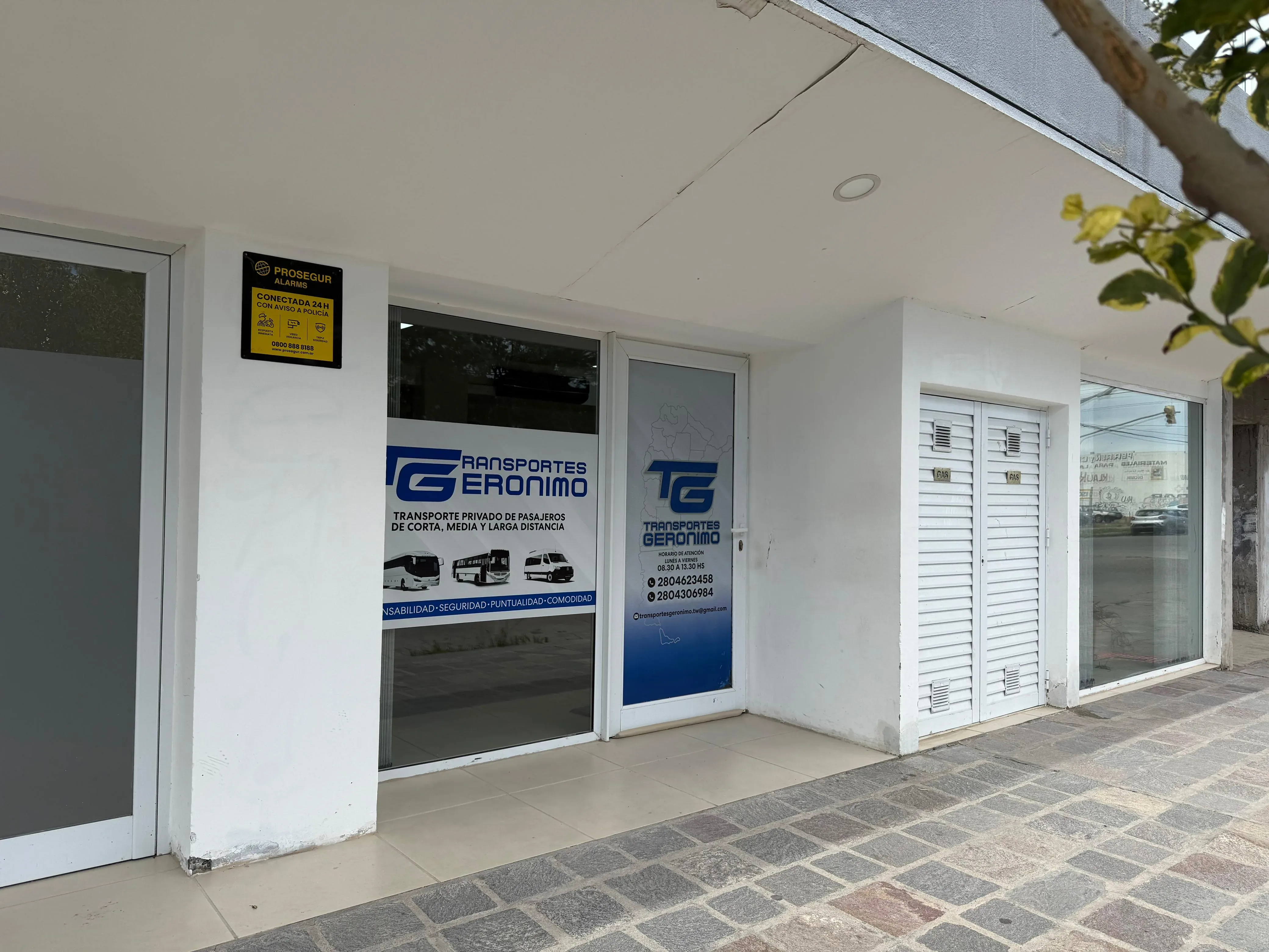The image shows the storefront of "Transportes Geronimo," featuring glass doors with company branding, a sign displaying various vehicle icons, and brick pavement outside the entrance.