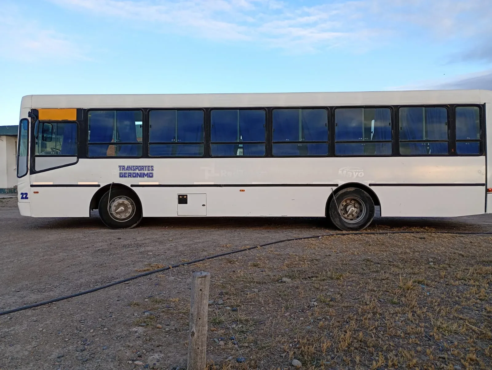 A white transit bus, labeled "Transportes Geronimo," is parked on a dirt road under a partly cloudy sky.