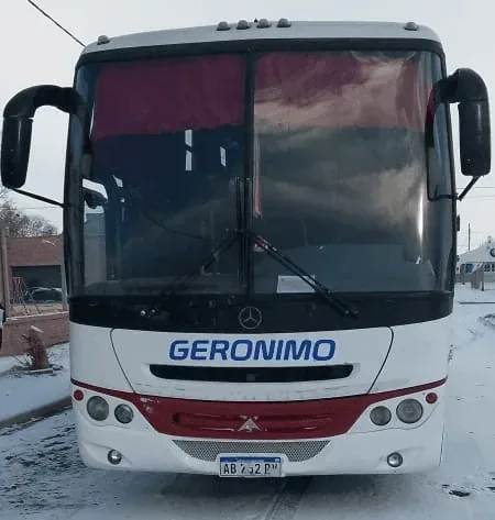 Front view of a white and red bus labeled "GERONIMO" parked on a snow-dusted road, featuring large windows, a Mercedes-Benz emblem, and a visible license plate.